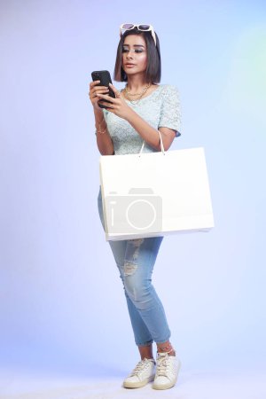 Photo for Full length portrait of a happy young woman holding shopping bag and mobile phone, isolated on a gradient blue background - Royalty Free Image