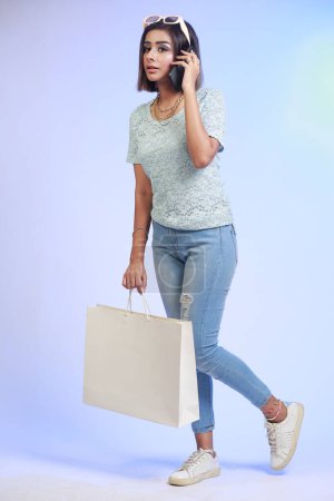 Foto de Retrato completo de una joven feliz sosteniendo una bolsa de compras y un teléfono móvil, aislado sobre un fondo azul degradado - Imagen libre de derechos