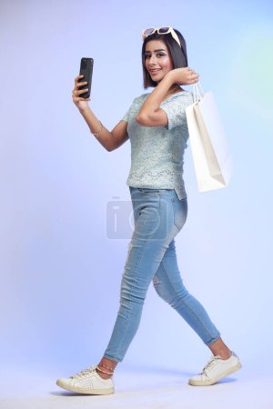 Téléchargez les photos : Portrait complet d'une jeune femme heureuse tenant son sac à provisions et son téléphone portable, isolée sur un fond bleu dégradé - en image libre de droit