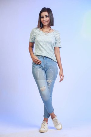Téléchargez les photos : Jeune fille indienne pakistanaise attrayante, en jeans et t-shirt, Poser et donner des expressions mignonnes sentiment sur un fond studio dégradé bleu - en image libre de droit