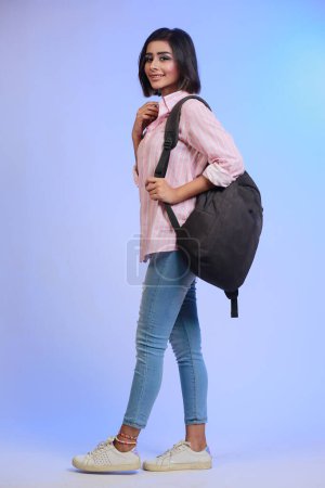 Téléchargez les photos : Photo de profil pleine longueur d'une étudiante en jeans et chemise rose marchant avec un sac à dos isolé sur fond de studio - en image libre de droit
