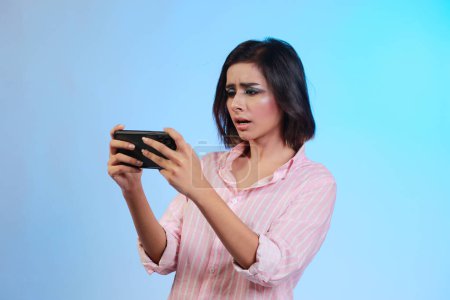 Foto de Foto de mujer joven positiva emocional posando aislado sobre azul gradiente pared fondo jugar juegos por teléfono móvil. - Imagen libre de derechos