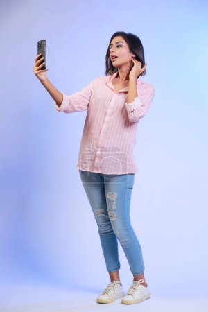 Téléchargez les photos : Image de belle fille hispanique prenant son selfie ou parlant sur un appel vidéo isolé sur fond bleu dégradé - en image libre de droit