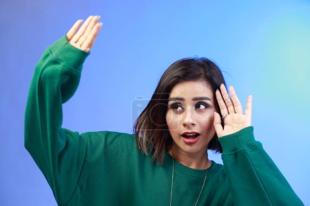 Photo for Side view portrait of scared female with raised arms, sees something awful, afraids, stop gesture, wearing green sweatshirt. Indoor studio shot isolated on gradient blue background - Royalty Free Image
