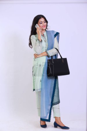 Photo for Pakistani woman descent, dressed in traditional attire, wearing a kamiz and shalwar. She has her hair open and is using a smartphone, as well as carrying a handbag. - Royalty Free Image