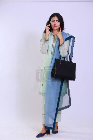 Photo for Pakistani woman descent, dressed in traditional attire, wearing a kamiz and shalwar. She has her hair open and is using a smartphone, as well as carrying a handbag. - Royalty Free Image