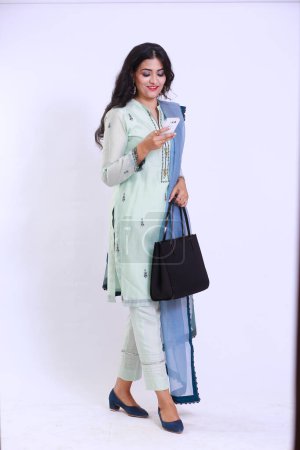Foto de Hermosa mujer paquistaní en bordado tradicional shalwar kameez vestido con dupatta. Caminar sosteniendo un smartphone con una sonrisa 'Desi Fashion Concept - Imagen libre de derechos