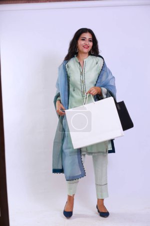 Foto de Young fashionable Pakistani woman walking after shopping with a paper bag looking in Camera. Image portrays the girl is happy with her purchase. Isolated over white background - Imagen libre de derechos