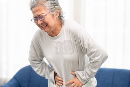 Photo for Senior woman suffering from stomach ache at home. Grabbing and squeezing belly with hand, feeling exhausted, standing in living room. Expressing pain on face - Royalty Free Image