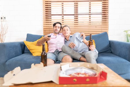 Photo for Two gay men in casual outfits sitting on sofa embracing and watching TV with beer and pizza in living room at home. Homosexual relationship concept. - Royalty Free Image
