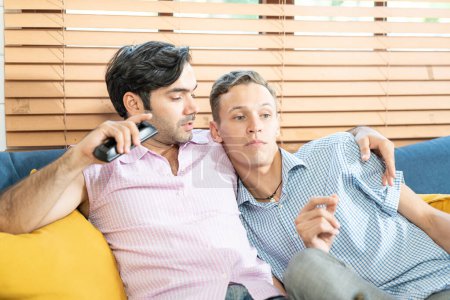 Photo for Two men in casual outfits sitting on sofa embracing and watching TV with beer and pizza in living room at home. LGBT gay relationship concept. - Royalty Free Image