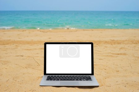 Téléchargez les photos : Ordinateur portable avec écran vierge pour un design créatif sur une plage de sable proche fond bleu de la mer. Ordinateur portable avec parcours de coupure de moniteur pour le modèle de maquette de conception de page de renvoi actuelle - en image libre de droit