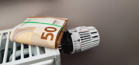 Bundle of euro banknotes on a heating radiator battery with thermostat temperature regulator. High costs for space heating and a significant increase in electricity prices in winter. Energy crisis and recession