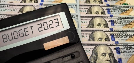 Budget in the new fiscal year 2023. Calculator with the words "budget 2023" on the screen against the background of a large number of bills of 100 us dollars