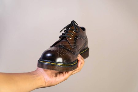 Die Hand eines Mannes hält einen dunkelbraunen Brogue-Schuh mit einer Gummisohle aus echtem Rindsleder. Männerhände mit eleganten und glänzenden Vintage-Schuhen auf beigem Hintergrund