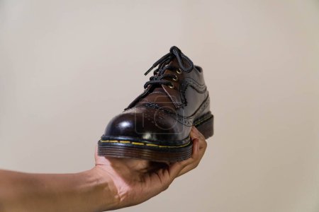 La mano de un hombre sostiene un zapato de punta de ala de brogue de degradado marrón oscuro con una suela de goma hecha de piel de vaca genuina. Manos de hombre sosteniendo zapatos vintage elegantes y brillantes sobre un fondo beige