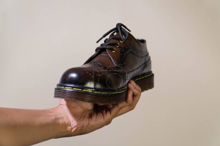 A man's hand holds a dark brown gradient brogue wingtip shoe with a rubber outsole made of genuine cowhide. Men's hands holding elegant and shiny vintage shoes on a beige background