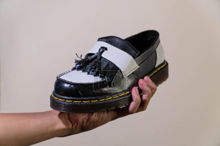 La mano de un hombre sostiene unos zapatos de borla en blanco y negro con una suela de goma hecha de piel de vaca genuina. Manos de los hombres sosteniendo elegantes y brillantes zapatos de dos tonos sobre un fondo crema