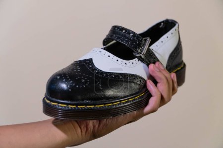 Die Hand eines Mannes hält einen schwarz-weißen Mary Jane Rockabilly Schuh mit Gummisohle aus echtem Rindsleder. Männerhände mit eleganten und glänzenden zweifarbigen Schuhen auf cremefarbenem Hintergrund