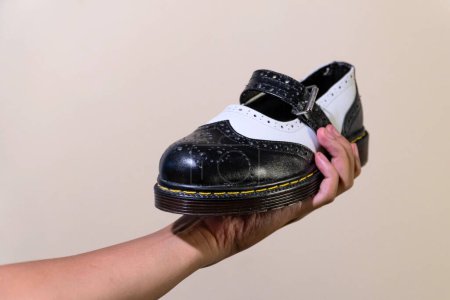 Die Hand eines Mannes hält einen schwarz-weißen Mary Jane Rockabilly Schuh mit Gummisohle aus echtem Rindsleder. Männerhände mit eleganten und glänzenden zweifarbigen Schuhen auf cremefarbenem Hintergrund