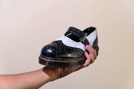 La mano de un hombre sostiene un zapato Mary Jane Rockabilly blanco y negro con una suela de goma hecha de piel de vaca genuina. Manos de los hombres sosteniendo elegantes y brillantes zapatos de dos tonos sobre un fondo crema