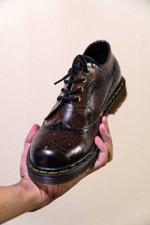 Die Hand eines Mannes hält einen dunkelbraunen Brogue-Schuh mit einer Gummisohle aus echtem Rindsleder. Männerhände mit eleganten und glänzenden Vintage-Schuhen auf beigem Hintergrund