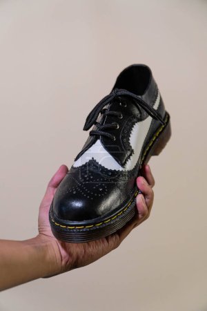 Die Hand eines Mannes hält einen schwarz-weißen Brogue-Schuh mit Gummisohle aus echtem Rindsleder. Männerhände mit eleganten und glänzenden zweifarbigen Schuhen auf cremefarbenem Hintergrund