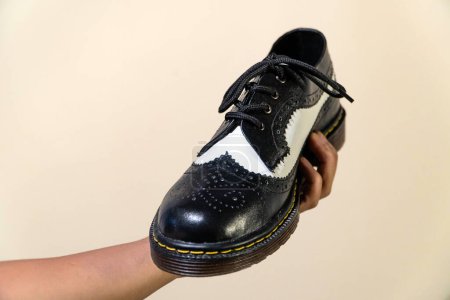 Die Hand eines Mannes hält einen schwarz-weißen Brogue-Schuh mit Gummisohle aus echtem Rindsleder. Männerhände mit eleganten und glänzenden zweifarbigen Schuhen auf cremefarbenem Hintergrund