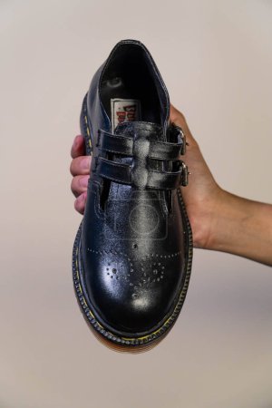 Die Hand eines Mannes hält einen schwarzen Mary Jane Rockabilly-Schuh mit Gummisohle aus echtem Rindsleder. Männerhände mit eleganten und glänzenden Vintage-Schuhen auf cremefarbenem Hintergrund