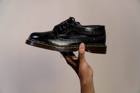 In der Hand hält ein Mann einen schwarzen Brogue-Schuh mit Gummisohle aus echtem Rindsleder. Männerhände, die elegante und glänzende Vintage-Schuhe auf cremefarbenem Hintergrund halten
