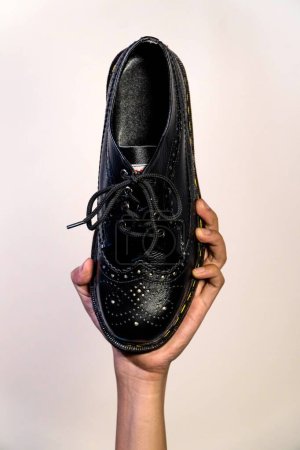 In der Hand hält ein Mann einen schwarzen Brogue-Schuh mit Gummisohle aus echtem Rindsleder. Männerhände, die elegante und glänzende Vintage-Schuhe auf cremefarbenem Hintergrund halten