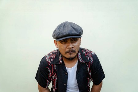 Macho man with beard and mustache looking serious isolated on beige background. Half body portrait of a vintage style Southeast Asian adult man in a patterned shirt, white tank top and newsboy cap