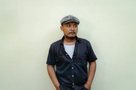 Close up of brutal macho man with beard and mustache looking serious and fierce isolated on beige background. Half body portrait of adult Southeast Asian man in black shirt and newsboy cap