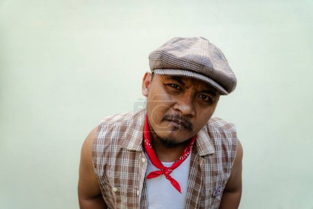 Southeast Asian macho man with beard and mustache looks serious and fierce isolated on beige background. Half body portrait of adult man in sleeveless shirt, bandana and newsboy hat