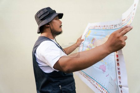 Asian adult man wearing travel outfit is looking at world map on beige background. Half body portrait of adult Southeast Asian man posing wearing vest and boonie hat reading a map