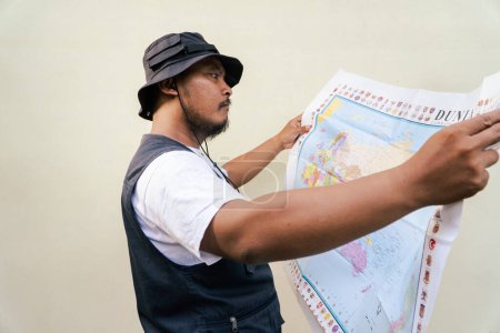 Hombre adulto asiático con traje de viaje está mirando el mapa del mundo sobre fondo beige. Retrato de medio cuerpo del hombre adulto del sudeste asiático posando con chaleco y sombrero boonie leyendo un mapa