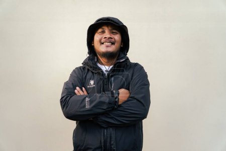 A macho adult man with a beard and mustache is posing seriously and smiling slightly against a cream background. Half body portrait of an adult Southeast Asian man wearing a hat, t-shirt and jacket