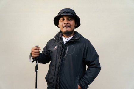 Portrait of Asian man wearing hiking outfit. Half body portrait of adult Asian man with beard and mustache posing climber wearing jacket, boonie hat and trekking poles isolated on beige background.