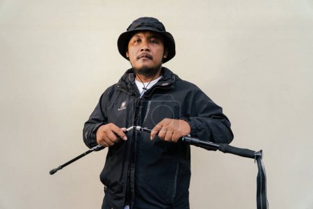 Portrait of Asian man wearing hiking outfit. Half body portrait of adult Asian man with beard and mustache posing climber wearing jacket, boonie hat and trekking poles isolated on beige background.