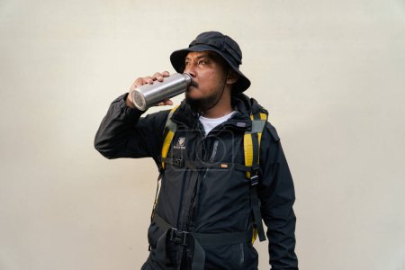 Mann im Reiseoutfit mit Jacke, Bommelmütze, Rucksack und Trinkflasche auf beigem Hintergrund. Halbkörperporträt eines erwachsenen asiatischen Mannes, der aus einem Fläschchen trinkend posiert