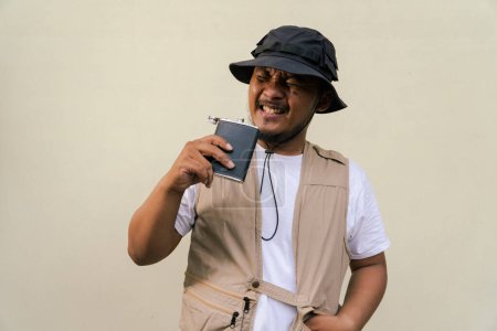 Mature asian man wearing travel outfit with vest, bucket hat and hip flask bottle isolated on beige background. Half body portrait of an adult Asian man posing drinking from a hip flask bottle