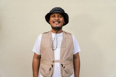 Macho mature man with beard and mustache wearing safari clothes isolated on beige background. Half body portrait of an adult Southeast Asian man striking various poses wearing a vest and bucket hat