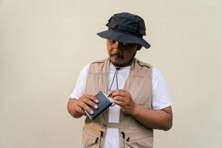 Mature asian man wearing travel outfit with vest, bucket hat and hip flask bottle isolated on beige background. Half body portrait of an adult Asian man posing drinking from a hip flask bottle