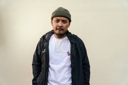 A macho mature man with a beard and mustache poses seriously and fiercely against a beige background. Half body portrait of adult Southeast Asian man posing wearing beanie cap, t-shirt and jacket