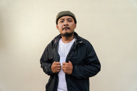 A macho mature man with a beard and mustache poses seriously and fiercely against a beige background. Half body portrait of adult Southeast Asian man posing wearing beanie cap, t-shirt and jacket
