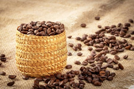 Photo for A cane basket filled with roasted coffee beans against the background of a burlap napkin. Coffee beans are scattered around - Royalty Free Image