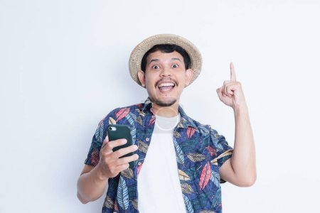 Photo for Asian man wearing beach shirt smiling happy while pointing finger up holding mobile phone - Royalty Free Image