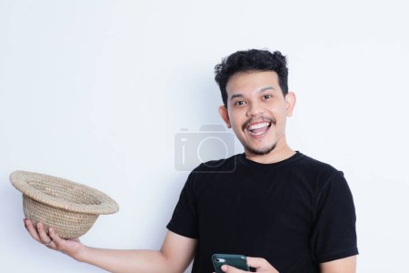 Foto de Hombre asiático con camisa negra casual y sombrero de playa sonriendo feliz mientras sostiene el teléfono móvil - Imagen libre de derechos