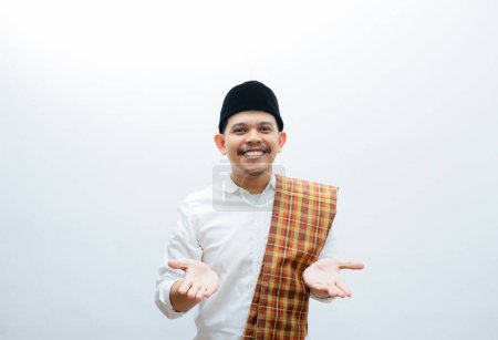 Asian Muslim man wearing white clothes  and sarung smiling to give greeting during Ramadan and Eid Al Fitr celebration standing over white background