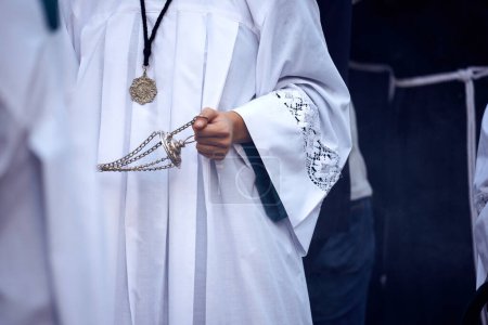 Photo for Detail of an altar boy in a white tunic in procession, holding a censer. He has a cross hanging around his neck. - Royalty Free Image